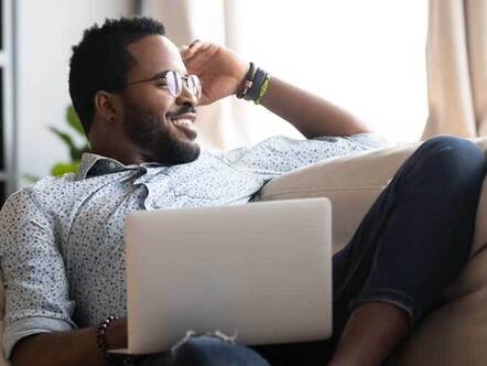 Smiling African American millennial man relax on comfortable couch in living room work on laptop gadget, happy biracial you